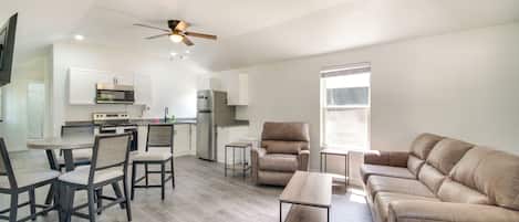 Un salon moderne comprenant un canapé, une table à manger avec des chaises, un coin cuisine équipé d’appareils électroménagers et un ventilateur de plafond.