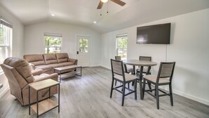 A spacious living room with a brown leather sofa, a wooden coffee table, and a dining area with a round table and four chairs. A ceiling fan is visible, and a flat-screen TV is mounted on the wall. - Walk to Lake: Cozy Grove Cabin w/ Fire Pit (Grove)