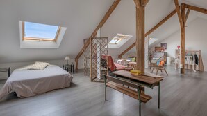 Une chambre spacieuse située dans les combles, avec de hauts plafonds, des fenêtres de toit, un grand lit, un banc en bois et un coin repas comprenant des chaises et une table.