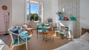 Un salon lumineux, meublé avec des meubles en osier, doté d’une cheminée et offrant une vue sur l’extérieur.