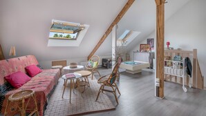 Une chambre à coucher confortable, aménagée en mezzanine, avec un plafond en pente, un lit recouvert d’une literie à motifs, une petite table ronde, des chaises en osier et une vue sur l’extérieur grâce à une lucarne.