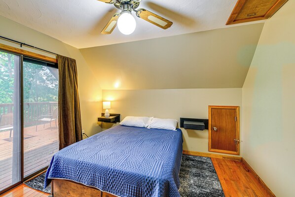 A bedroom with a bed, a ceiling fan, a nightstand, a lamp, a wardrobe, and a sliding glass door leading to an outdoor deck.