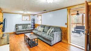 A living room with a grey sofa, a coffee table, and a television. There is a wooden floor and a door leading to an outdoor area with a bench. - Peaceful South Holston Lake Cabin w/ Dock & Deck! (Abingdon)