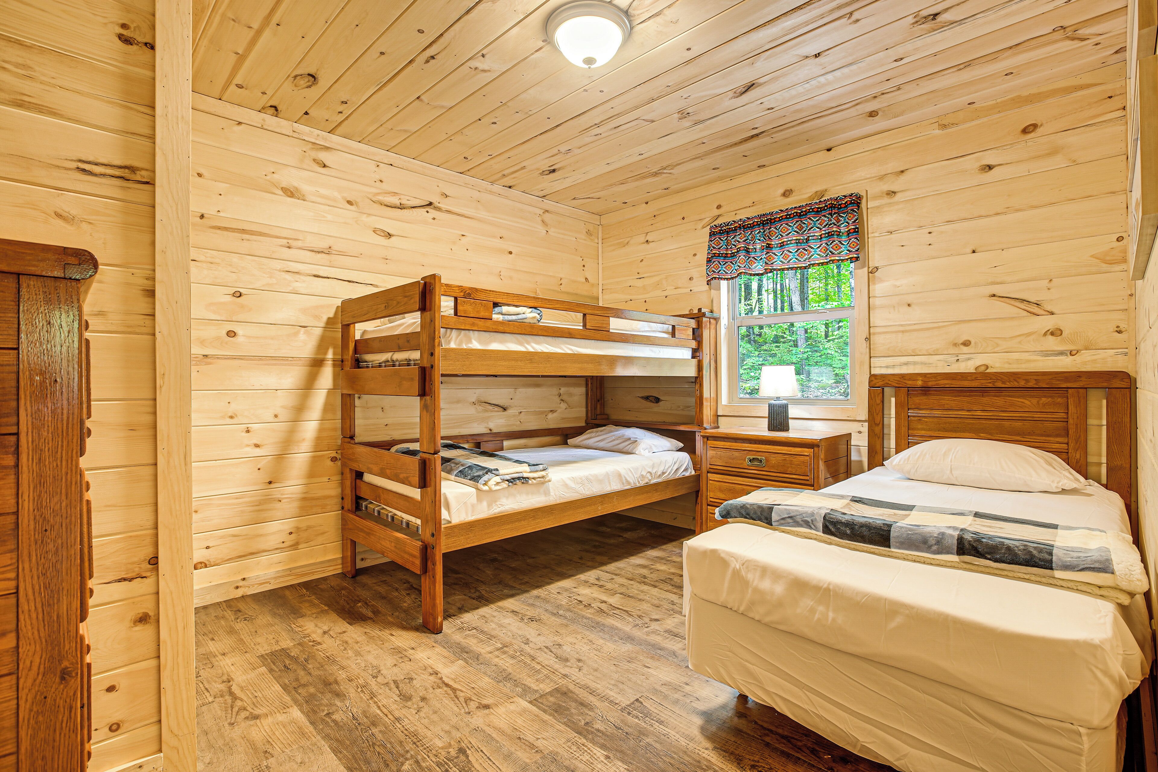 A wooden cabin room with bunk beds, a single bed, a nightstand, and a window with a view of greenery.