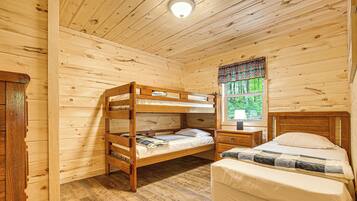 A wooden cabin room with bunk beds, a single bed, a nightstand, and a window with a view of greenery.