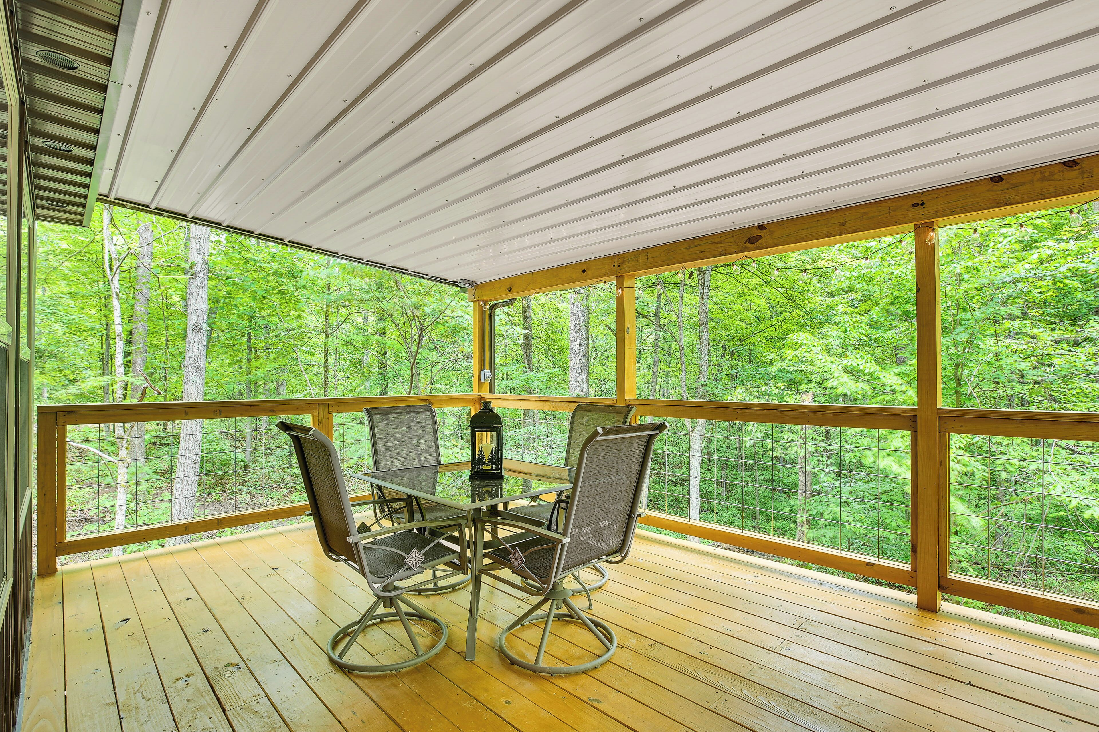 A wooden deck with a table and chairs, surrounded by glass railings, overlooking a forest.
