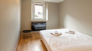 Une petite chambre bien éclairée, avec un lit simple, une petite table ronde et une fenêtre donnant sur l’extérieur.