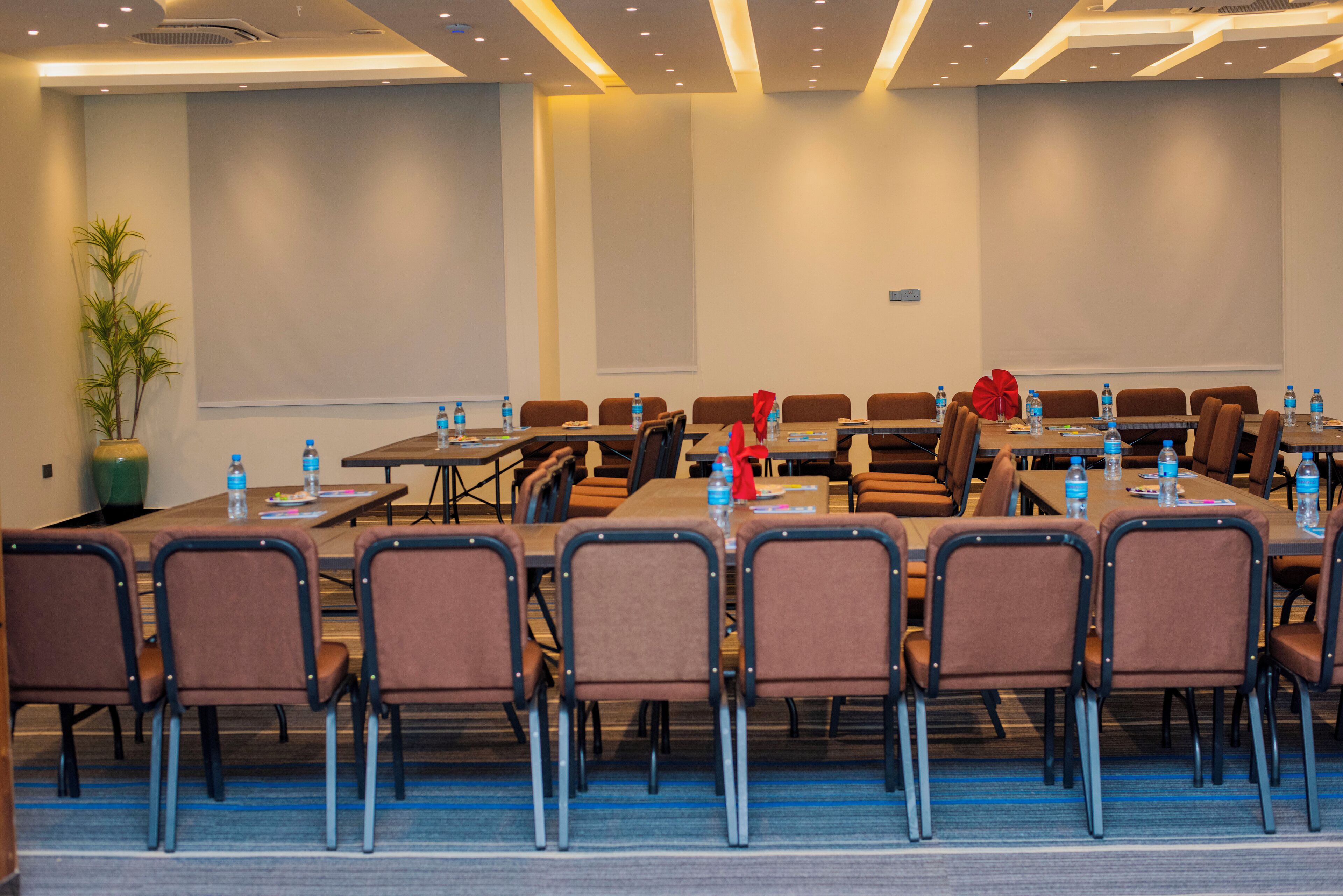 Une salle de conférence équipée de plusieurs tables et chaises, avec des bouteilles d’eau sur chaque table et un écran de projection au mur.