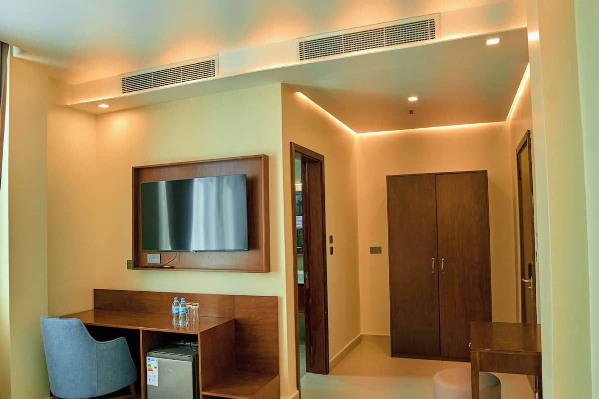 A hotel room with a bed, a desk with a computer, a TV mounted on the wall, a wardrobe, and a small ottoman.