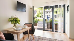 A modern living room with a dining table, a sofa, a TV mounted on the wall, and a sliding glass door leading to a garden.