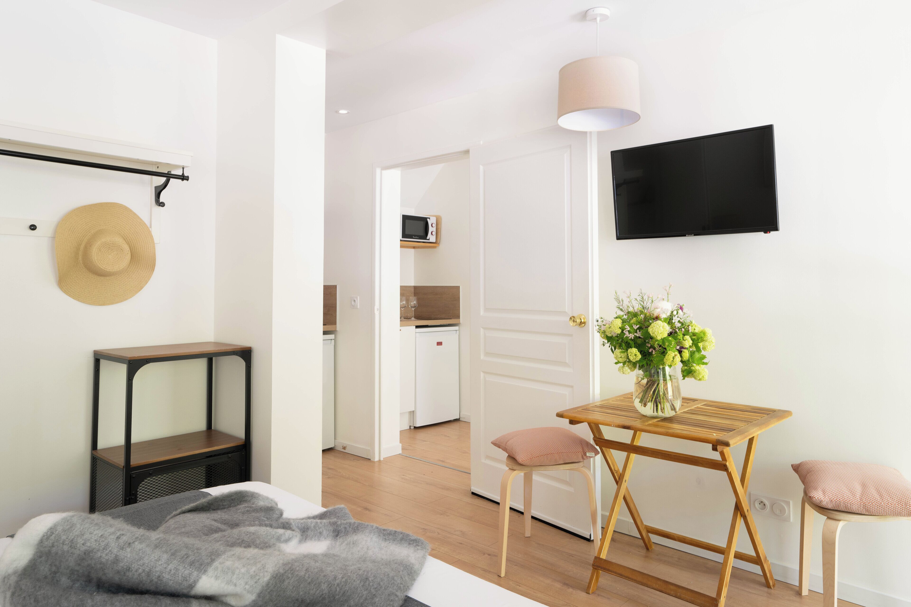 A modern, well-lit room with a bed, bedside table, and a small table with chairs. A TV is mounted on the wall, and a door leads to a kitchen area.