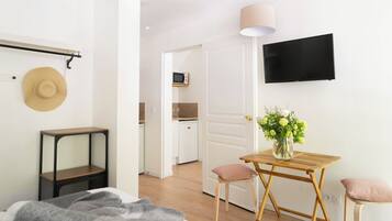 A modern, well-lit room with a bed, bedside table, and a small table with chairs. A TV is mounted on the wall, and a door leads to a kitchen area.