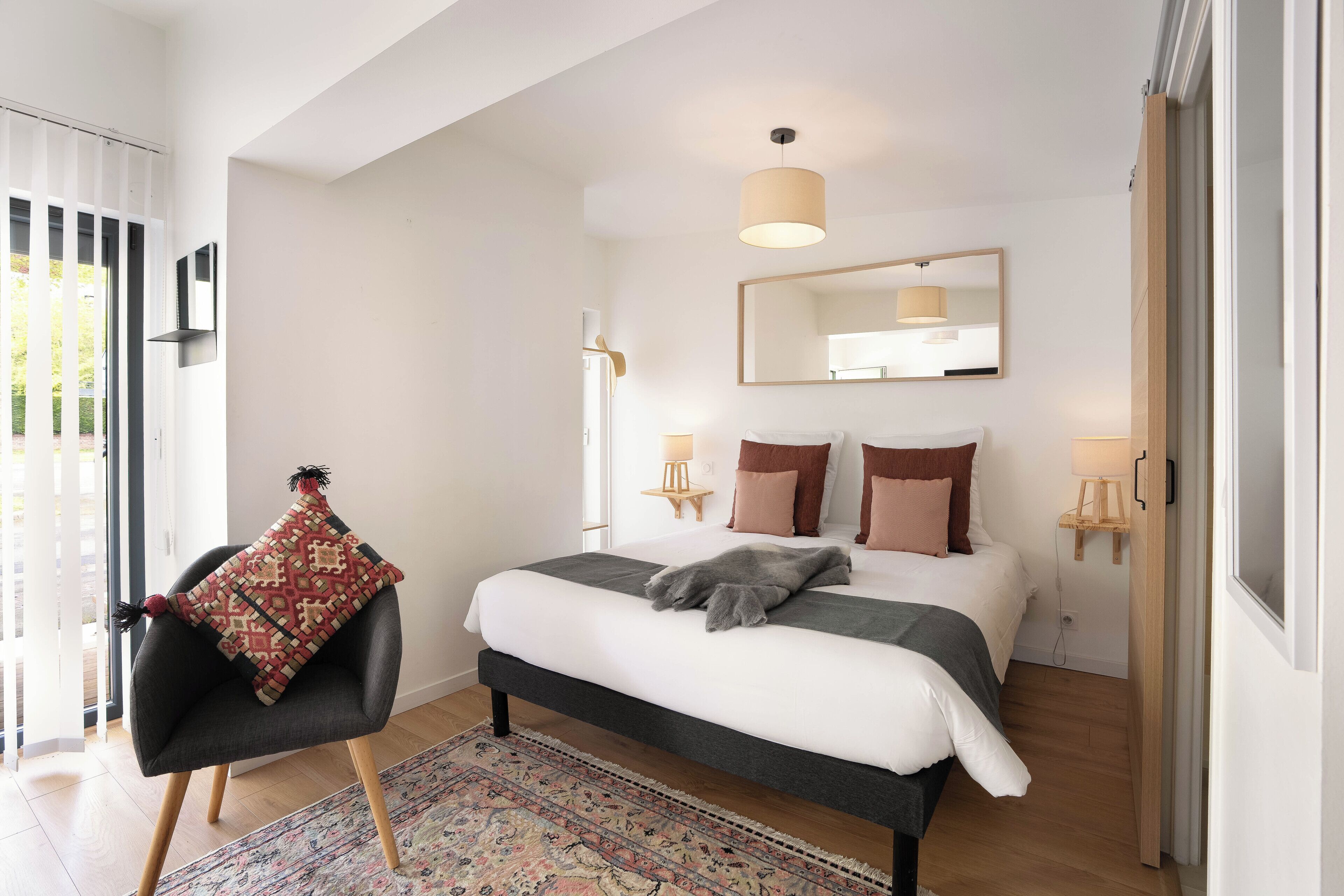 Un dormitorio moderno con una cama, un sillón con cojín estampado, un espejo grande y una lámpara colgante.