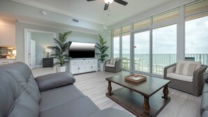 Un salon spacieux avec un canapé gris, une table basse en bois et des fauteuils en osier. Un téléviseur à écran plat est fixé sur un meuble blanc, et de grandes fenêtres offrent une vue sur le balcon.