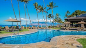 A swimming pool with lounge chairs and umbrellas, surrounded by palm trees, with a mountain in the background. - Wailea Ekahi 37e 1 Bedroom Condo (Kihei)