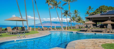 A swimming pool with lounge chairs and umbrellas, surrounded by palm trees, with a mountain in the background.