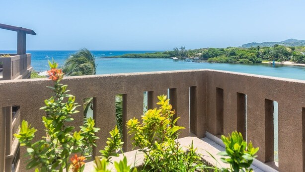 A balcony with a view of a calm sea, a pier, and lush greenery. - Pelican Penthouse -mangrove Dock 3 Bedroom Home by RedAwning (Roatan)