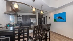 A modern kitchen with dark cabinets, a stainless steel refrigerator, and a large island with bar stools. There is a painting of a turtle on the wall. - Pelican Penthouse -mangrove Dock 3 Bedroom Home by RedAwning (Roatan)