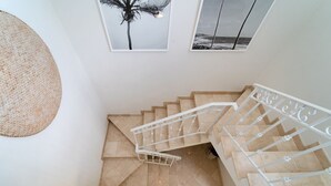Un escalier aux carreaux beiges et aux rampes blanches, menant à un palier où sont accrochées des photos encadrées représentant un palmier et une scène de plage.