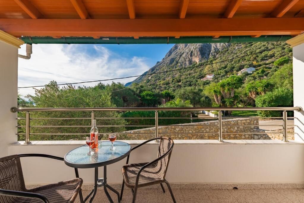 Un balcón con mesa y sillas, con vista a un paisaje montañoso con vegetación y un sendero de piedra.