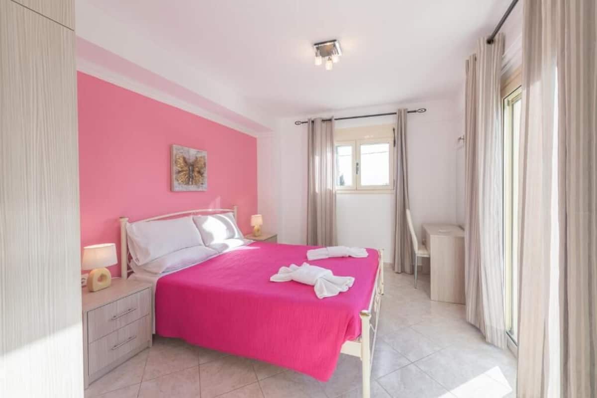 A bedroom with a pink wall, a bed with white bedding, a bedside table with a lamp, and a window with curtains.