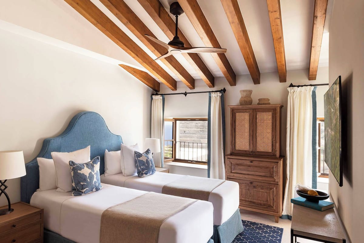 A hotel room with two beds, wooden beams on the ceiling, a window with curtains, a bedside table with a lamp, and a cabinet.
