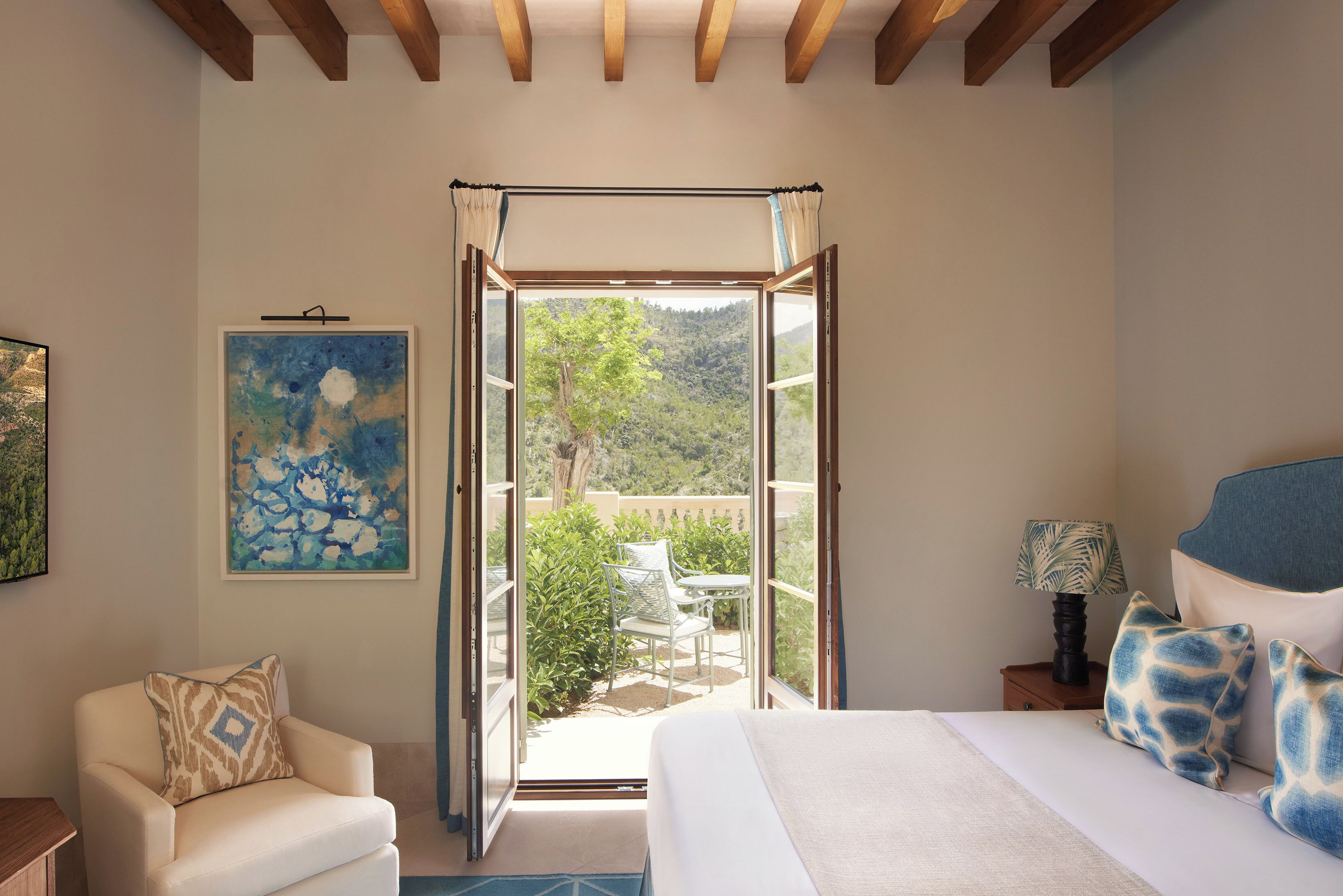Une chambre à coucher comprenant un lit, une chaise, une table de chevet, une lampe, un tableau et une porte ouverte donnant sur un balcon avec vue sur la verdure.