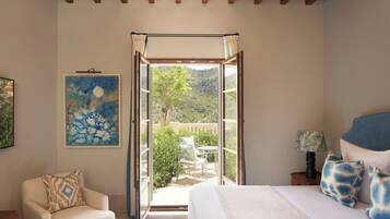 Une chambre à coucher comprenant un lit, une chaise, une table de chevet, une lampe, un tableau et une porte ouverte donnant sur un balcon avec vue sur la verdure.