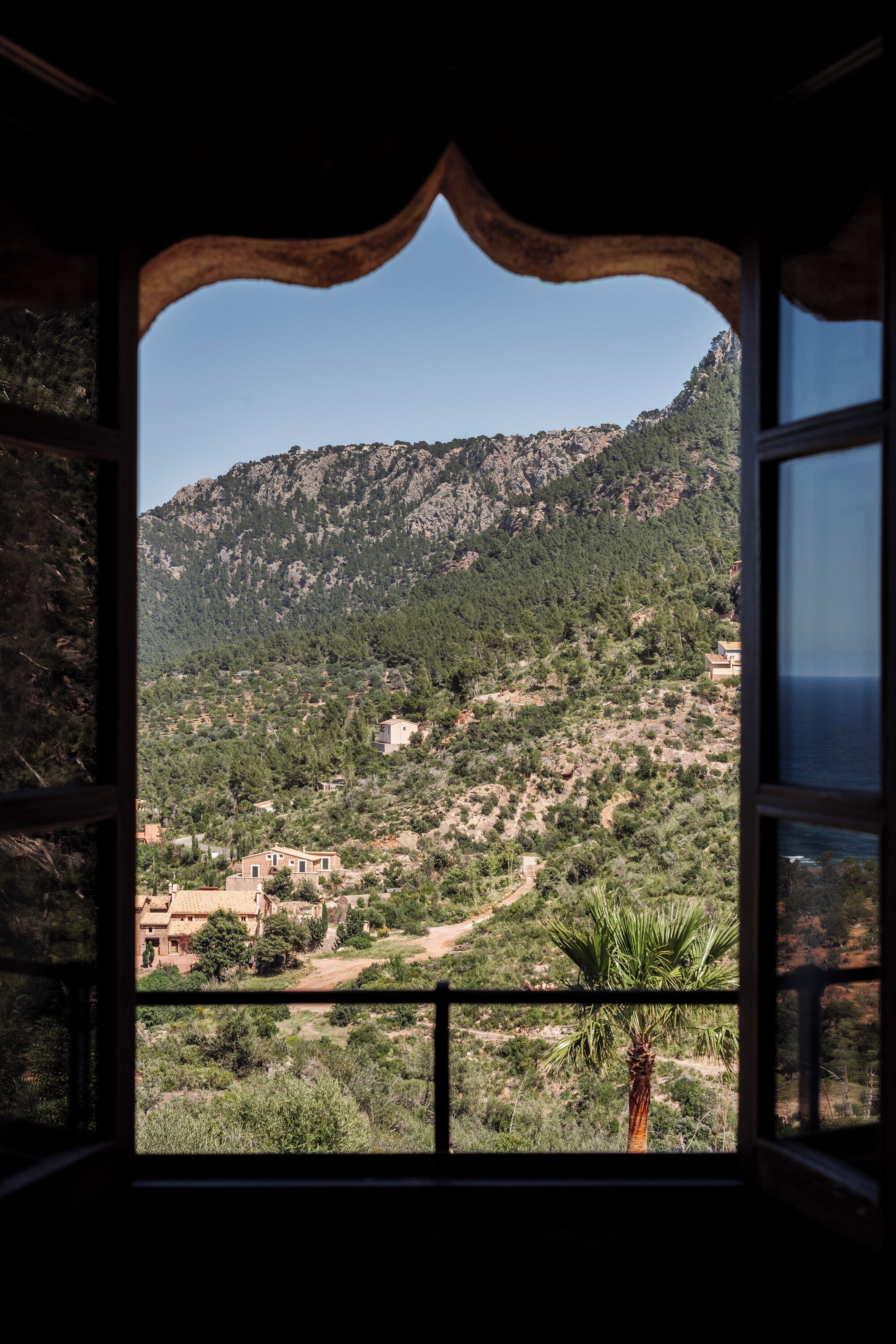 Vista a través de un ventanal arqueado de un paisaje montañoso con edificios dispersos y una palmera.