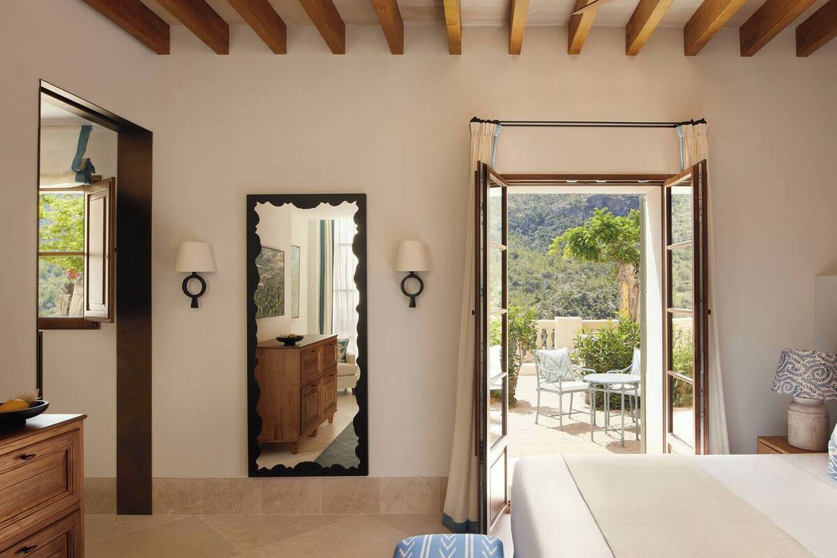 A bedroom with a large bed, a wooden dresser, a mirror, and a view of an outdoor patio with chairs and a table.