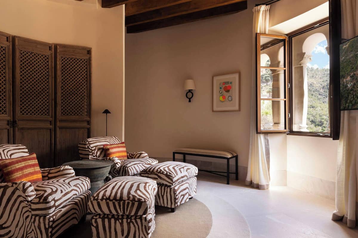 A cozy room with a seating area featuring striped armchairs and ottomans, a small table, and a window with a view of greenery.
