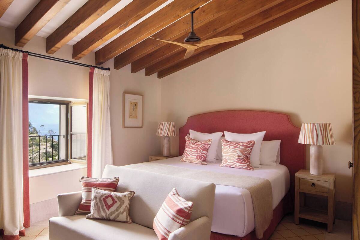 A bedroom with a large bed, a chair, a nightstand, a window with curtains, and a ceiling fan.