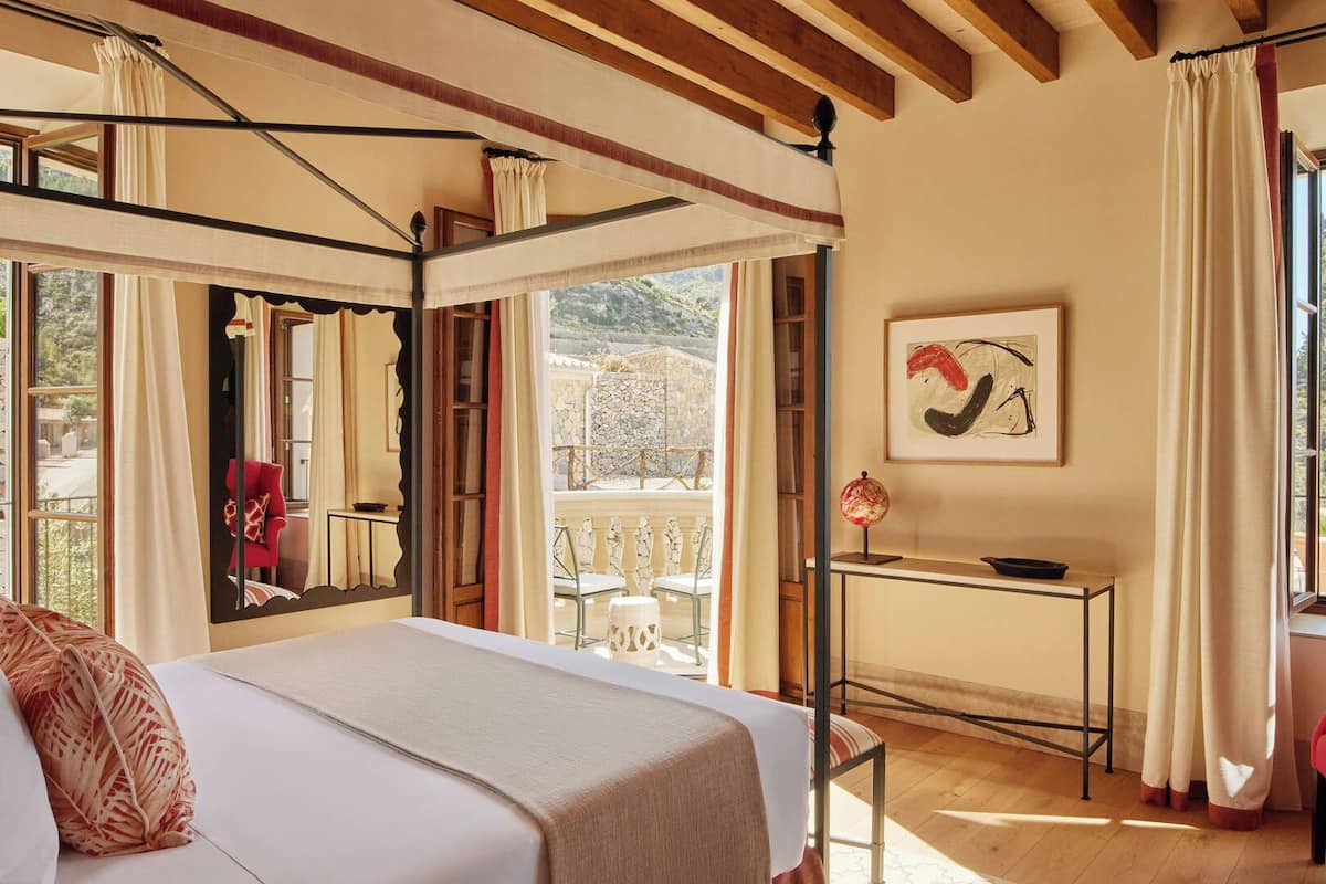 A spacious bedroom with a large bed, a desk, and a chair. There is a balcony with a view of the landscape.