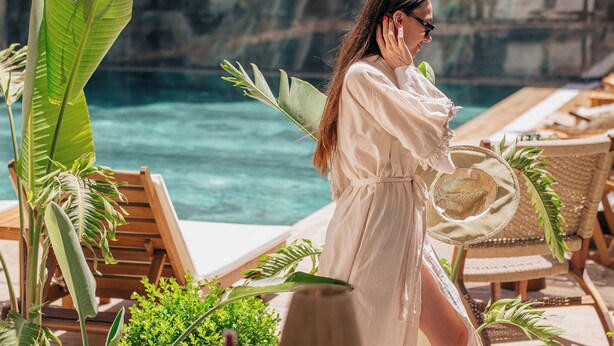 A person in a white dress walking by a pool with wooden lounge chairs and tropical plants. - Los Suites (Konyaalti)