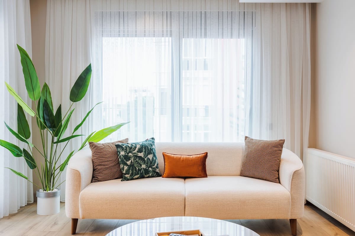 Sala de estar com sofá bege, duas almofadas estampadas, uma planta verde grande e um radiador branco.
