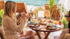 A dining table with a variety of foods and drinks, surrounded by wooden chairs, set against a backdrop of a body of water and greenery. - Los Suites (Konyaalti)