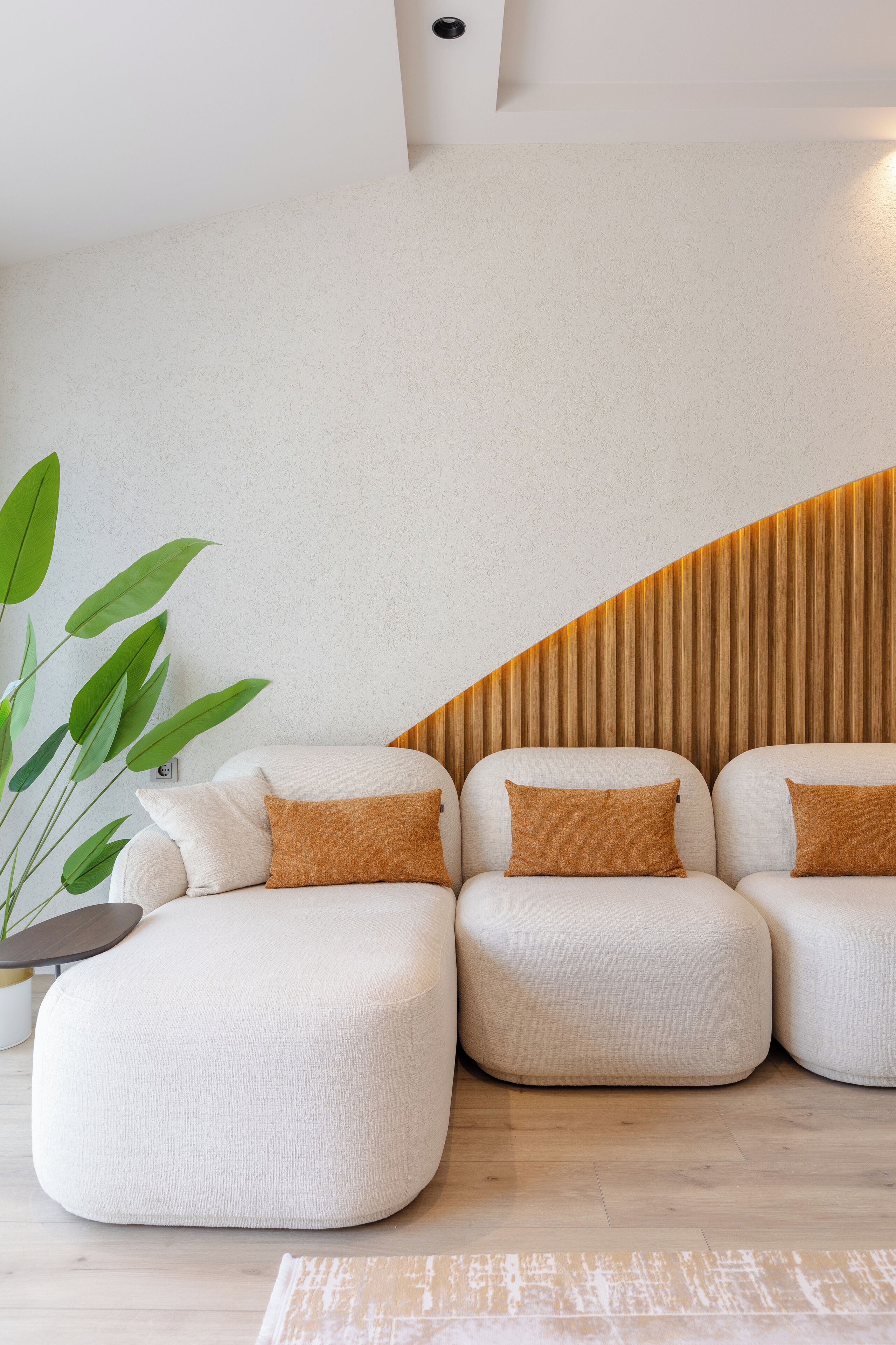 Een moderne woonkamer met een hoekbank, een grote groene plant en een wand van houten latten.
