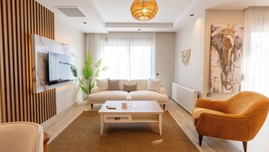 A modern living room with a sofa, coffee table, and armchair, a television mounted on the wall, and a large window with curtains. - Los Suites (Konyaalti)