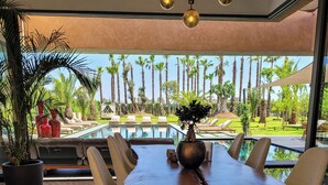 Comedor con una gran mesa, sillas y vistas a una piscina y palmeras.