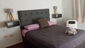 Ein ordentlich gemachtes Bett mit grauem Kopfteil, pinken und braunen Kissen und einem weißen Nachttisch mit Lampe.