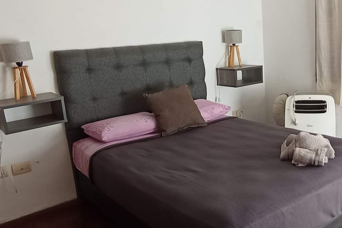 Ein ordentlich gemachtes Bett mit grauem Kopfteil, pinken und braunen Kissen und einem weißen Nachttisch mit Lampe.