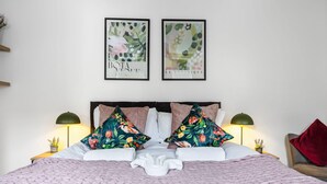 Un lit bien fait, recouvert d’une couverture rose, avec deux coussins à motifs floraux et une serviette blanche au centre. Deux lampes murales sont fixées de part et d’autre du lit.
