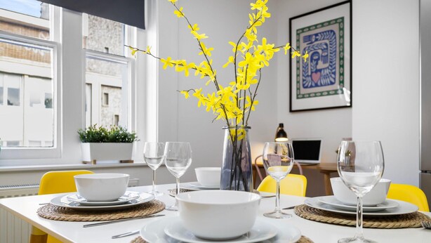 Une table à manger dressée avec des bols, des assiettes et des verres à vin blancs, un vase rempli de fleurs jaunes et des chaises jaunes.