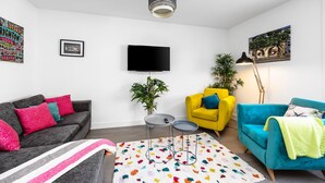 Un salon avec un canapé gris, un fauteuil jaune, un fauteuil bleu, un téléviseur fixé au mur et une lampe sur pied.