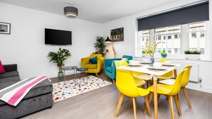 Un salon moderne avec un canapé gris, un fauteuil jaune, un canapé bleu, une table à manger avec des chaises jaunes et un téléviseur à écran plat fixé au mur.