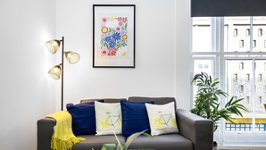 A modern living room with a sofa, decorative pillows, a floor lamp, a framed artwork, and a view of buildings through a window. - Queens Mile 1 With Free Parking By My Getaways (Brighton)