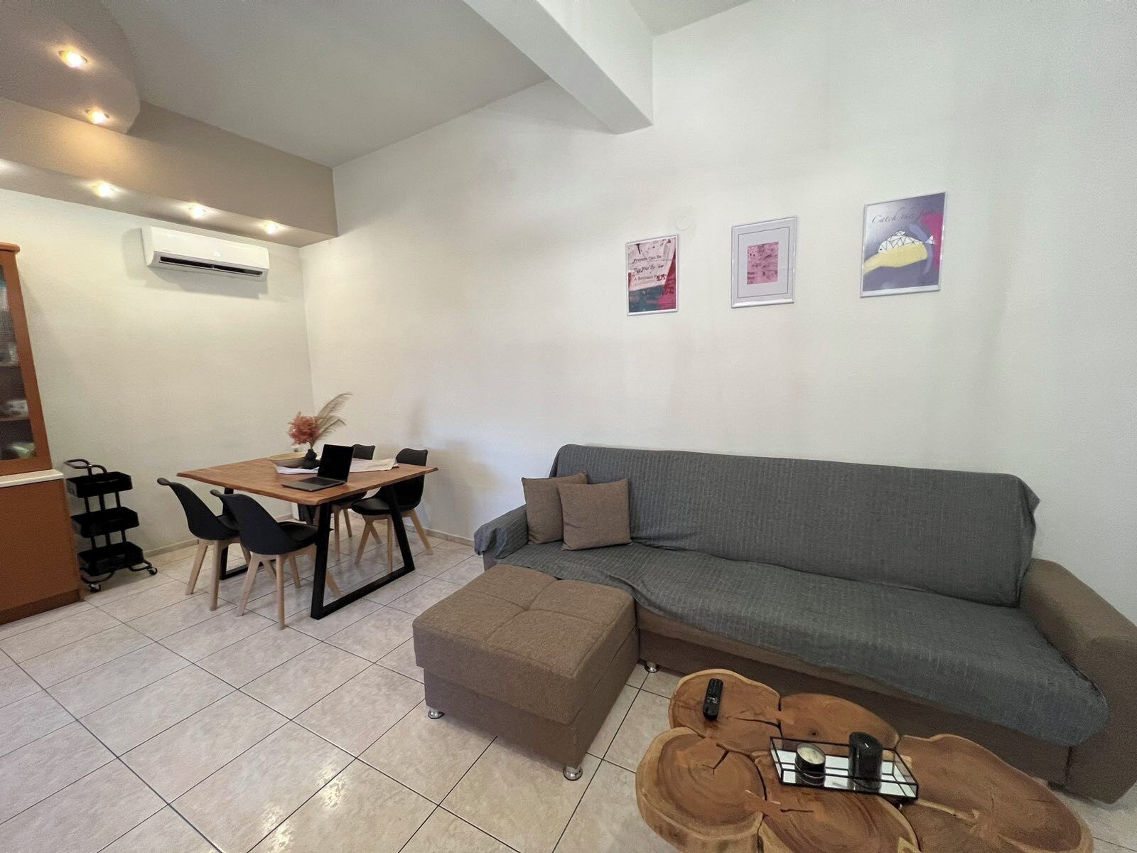 A modern living room with a grey sofa, a wooden dining table with chairs, a small ottoman, and a coffee table. There are two framed pictures on the wall.