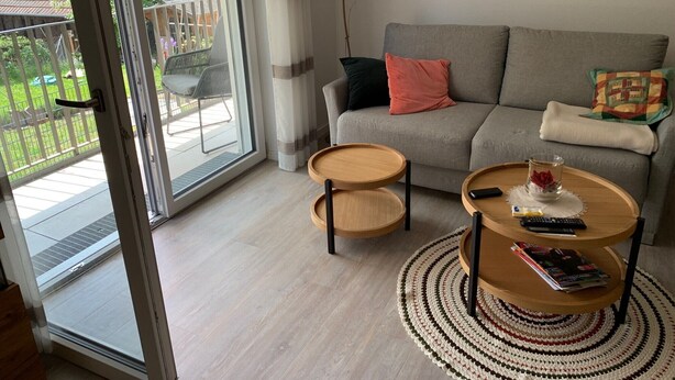 A modern living room with a grey sofa, two side tables, a coffee table, and a view of a balcony with outdoor furniture.