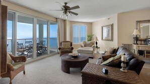 A spacious living room with a balcony overlooking the sea, furnished with wicker furniture, a ceiling fan, and a large window with shutters. - Soak Up The Sun - Beach Colony East PH 17C (Navarre)