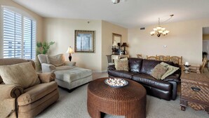 A living room with a brown leather sofa, a beige armchair, a wicker ottoman, and a dining area with a wooden table and chairs. - Soak Up The Sun - Beach Colony East PH 17C (Navarre)